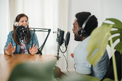 Content multiracial male and female coworkers speaking in mics while looking at each other and recoding podcast in modern studio